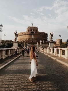 a woman in white dress walking on brick walkway next to large building with statues around it