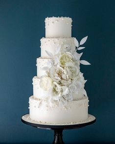 a three tiered wedding cake with white flowers on top and blue wall in the background