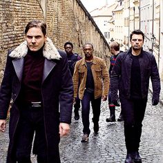 a group of men walking down a cobblestone street in an alley way with one man wearing a fur collar