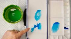 a person is painting with blue and green paints on a white tray next to a green cup