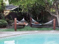 two hammocks sitting next to a swimming pool in a backyard with trees and bushes