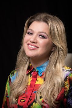 a woman with long blonde hair wearing a colorful shirt and smiling at the camera in front of a black background