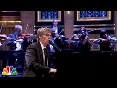 a man in a suit and tie playing the piano while other people are on stage behind him