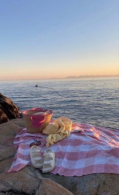 a towel, basket and sandals are sitting on the rocks by the water at sunset