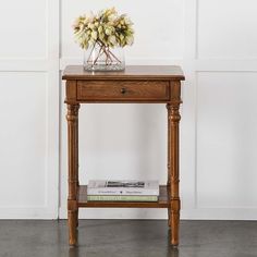 a small wooden table with a flower arrangement on top and magazines under it, against a white wall