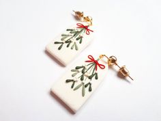 two white ceramic earrings with red bows and green leaves on them, hanging from gold - plated hooks