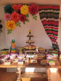 a table topped with lots of cakes and cupcakes next to a wall covered in paper flowers