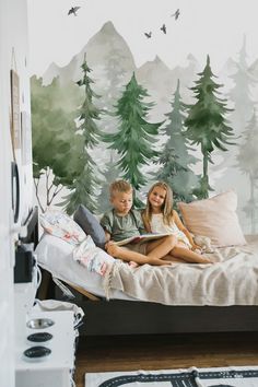 two children sitting on a bed in front of a forest mural