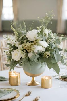 a table with white flowers and greenery in a gold vase on top of it