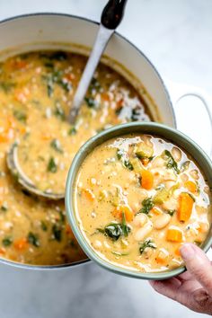 a person holding a bowl of soup with spoons in it and another pot full of soup on the side