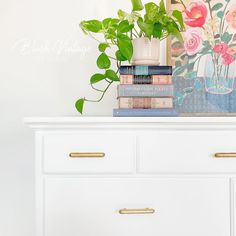 a white dresser topped with lots of books next to a vase filled with pink flowers