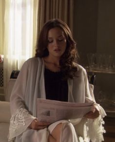 a woman sitting on top of a white couch next to a window holding a newspaper