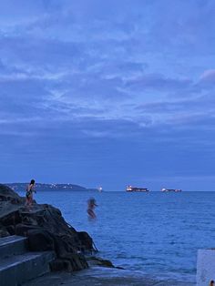 two people are standing on the rocks near the water at dusk, one person is flying a kite