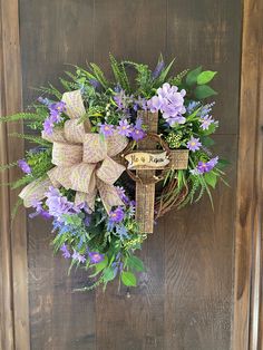 a wreath with purple flowers and greenery hanging on a wooden door, decorated with a cross