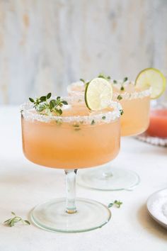 two margaritas sitting on top of a table with lime slices and garnish