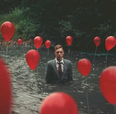 a man in a suit and tie standing in water with red balloons floating around him