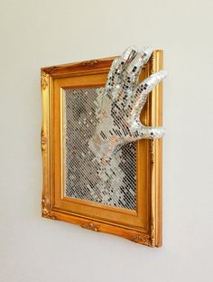 a gold frame with a hand made out of silver sequins hanging on the wall