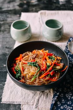 a bowl filled with noodles and vegetables on top of a table next to two cups