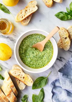 basil pesto in a white bowl surrounded by bread and lemons on a marble surface