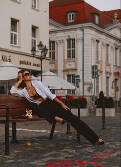 a woman sitting on top of a wooden bench next to tall buildings and wearing sunglasses