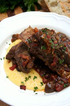 a white plate topped with meat and grits next to bread on top of a wooden table