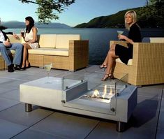 three people sitting on wicker couches around an outdoor fire pit with water in the background