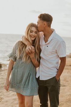 a man and woman standing on top of a sandy beach next to the ocean with their arms around each other