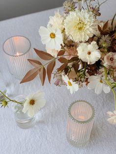 flowers and candles are sitting on a white tablecloth with two glass vases filled with flowers