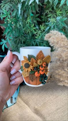 a person holding a coffee cup with autumn decorations on it and a teddy bear in the background