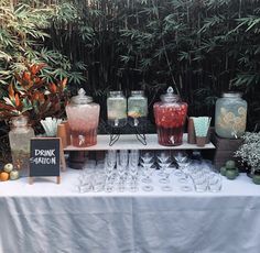 a table topped with lots of glasses and jars filled with liquid next to plants in front of a bamboo fence