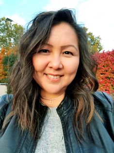 a woman in a leather jacket is smiling for the camera with trees behind her and red leaves on the ground