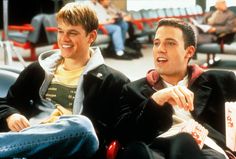 two young men sitting next to each other at an airport, one holding a drink and the other eating popcorn