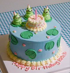 a birthday cake decorated with three little green frogs sitting on top of each other in front of a blue checkered tablecloth