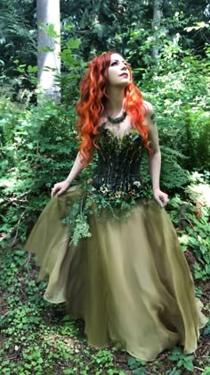 a woman with red hair and green dress standing in the woods looking up into the sky