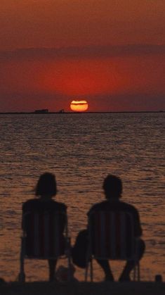 two people sitting on lawn chairs watching the sun go down over the ocean in silhouette