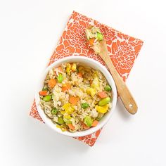 a bowl filled with rice and vegetables next to a wooden spoon on an orange napkin