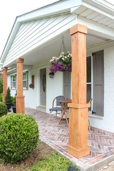 a porch covered in plants and flowers next to a white brick building with wooden pillars