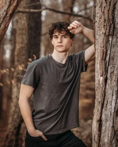 a young man standing next to a tall tree in the woods with his hands on his head