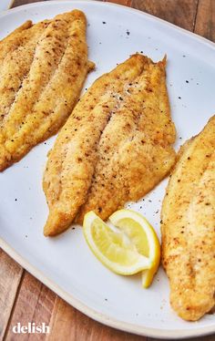 three fish fillets on a white plate with lemon wedges