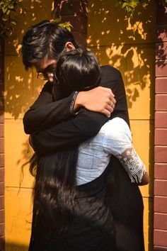 two people hugging each other in front of a yellow brick wall and green leaves on the tree