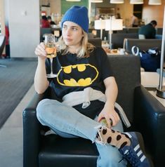 a woman sitting in a chair holding a glass of wine and wearing a batman t - shirt