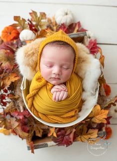 a newborn baby wrapped in a yellow blanket and wearing a knitted hat is laying on a white bowl surrounded by autumn leaves