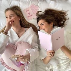 two women laying in bed reading books and smiling at each other with pink sticky notes on the wall behind them