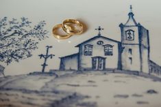 two gold wedding rings sitting on top of a blue and white plate with a drawing of a church