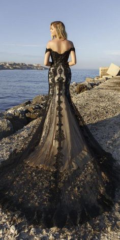 a woman in a black dress standing on the rocks by the water with her back to the camera