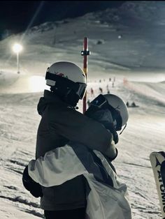 two skiers hugging each other on the snow covered slope at night with lights in background