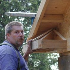 a man holding a piece of wood up to the top of a wooden structure in front of trees