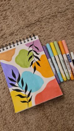 a notebook with markers and pens on the floor next to it are four different colored leaves