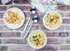 three bowls of soup and two spoons on a brick wall with floral table cloth