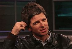 a man in a black leather jacket is talking on the set of an interview with his hand up to his ear